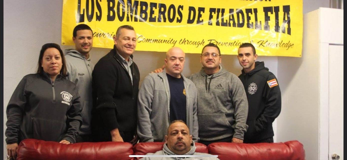 Group portrait of SAPFA members in front of the Los Bomberos de Filadelfia banner — Fuego y Vida (Fire and Life)