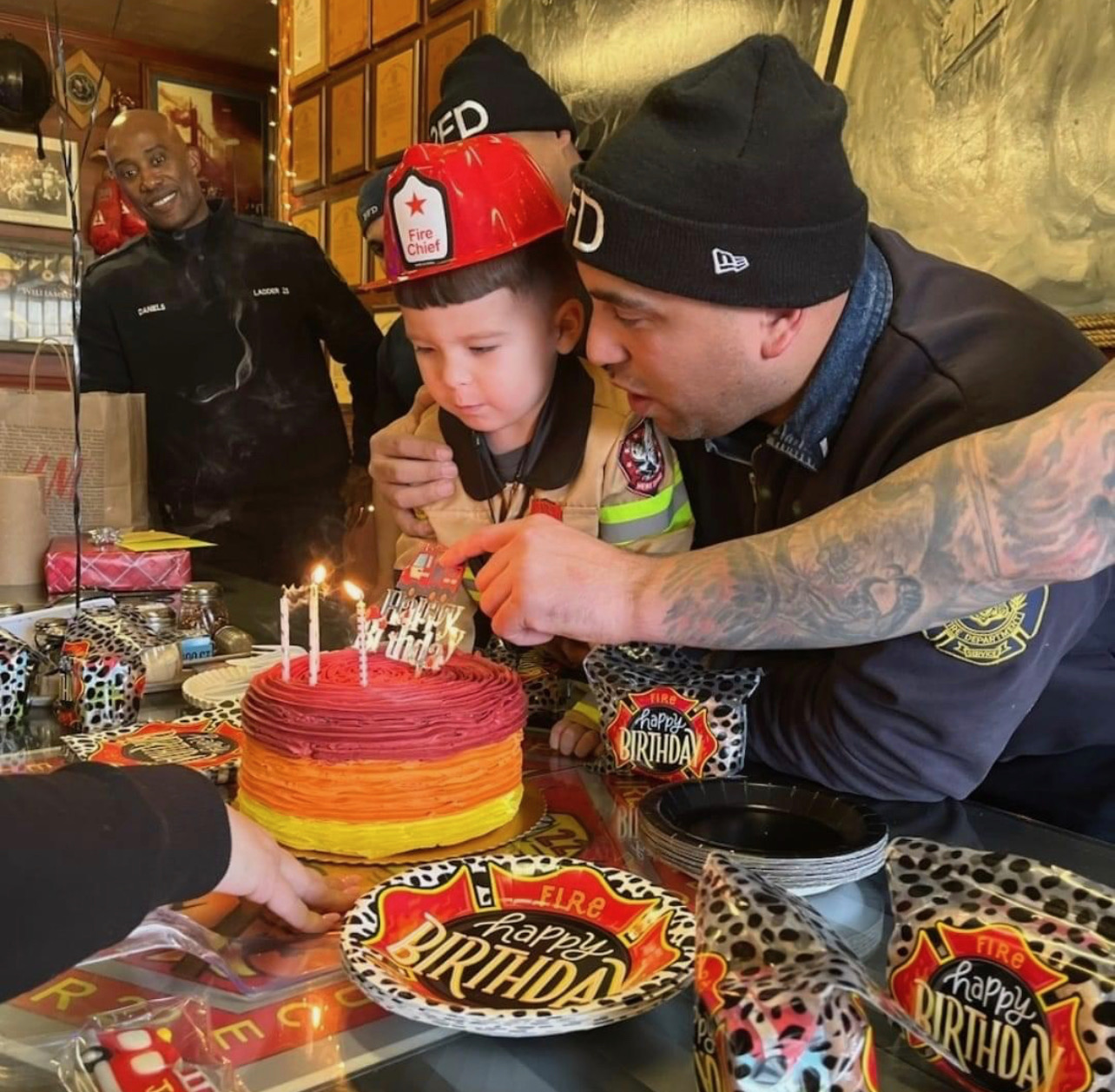 A young child wearing a fire chief helmet blowing out birthday candles with firefighters around him