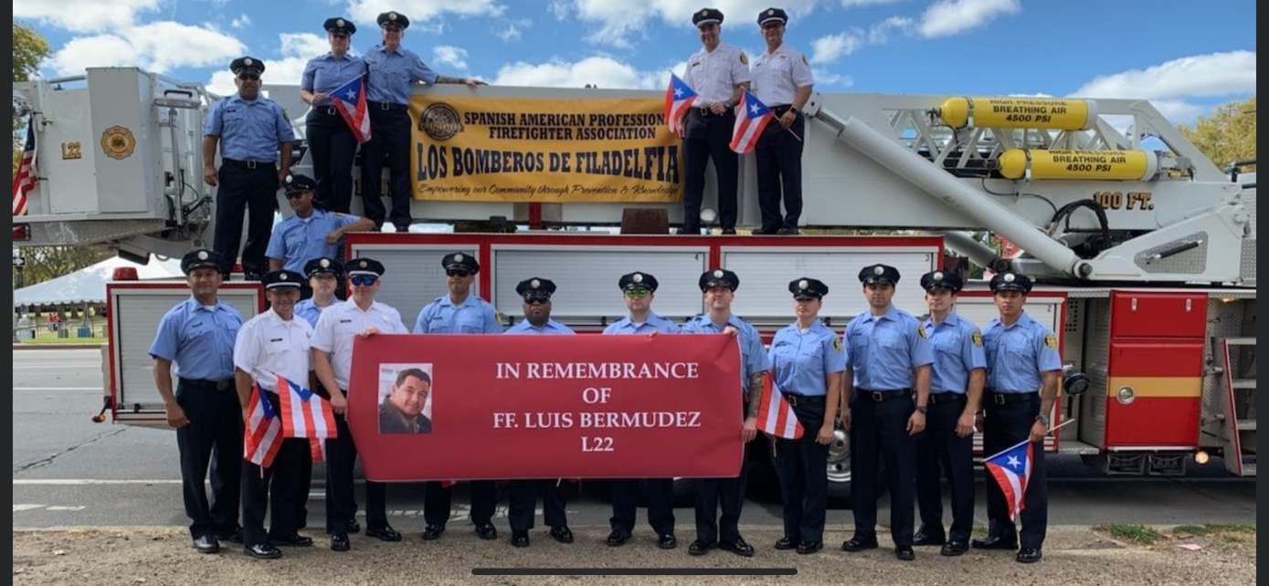 SAPFA members on a fire truck with the Los Bomberos de Filadelfia banner and a remembrance banner for FF. Luis Bermudez, L22